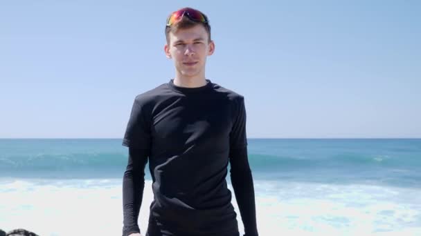 Jonge fit aantrekkelijke knappe man in het zwart tonen duimen tot aan de camera staande op het strand. Sterke golven raken het strand op de achtergrond. — Stockvideo