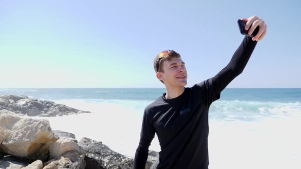 Młody szczęśliwy uśmiechnięty człowiek rozmawia Zdjęcia z niego śmiać się i pokazując kciuki się na skalistej plaży z fale rozpryskiwania znowu skały. Zwolnionym — Wideo stockowe