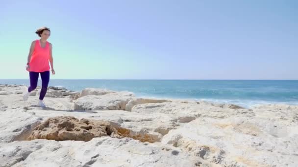 Giovane ragazza caucasica attraente che corre sopra le rocce vicino al mare. Onde che colpiscono la spiaggia rocciosa e spruzzi d'acqua . — Video Stock