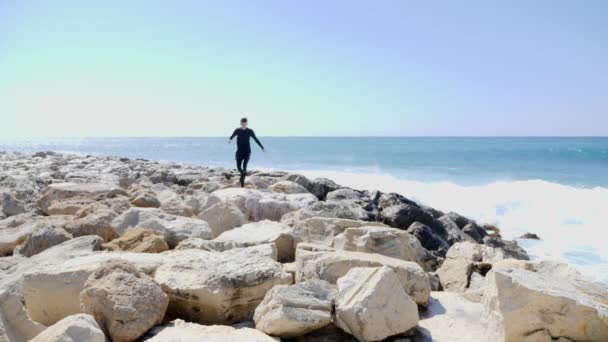 Desportista jovem em forma correndo e pulando sobre as rochas em uma praia com fortes ondas oceânicas atingindo as falésias e salpicos de água. Movimento lento — Vídeo de Stock