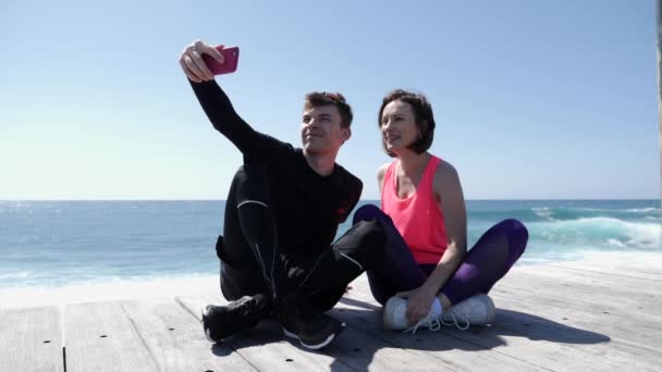 Pareja joven en forma sentada cerca de la playa sosteniendo el teléfono y tomando selfies posando. Feliz novio y novia tomando fotos. Moción lenta — Vídeo de stock