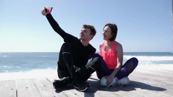 Pareja joven en forma sentada cerca de la playa sosteniendo el teléfono y tomando selfies posando. Feliz novio y novia tomando fotos — Vídeos de Stock