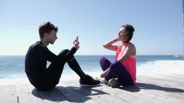 Jovem homem apto sentado na praia tirando fotos de sua namorada com o telefone. Ondas fortes salpicando againts as rochas no fundo — Vídeo de Stock
