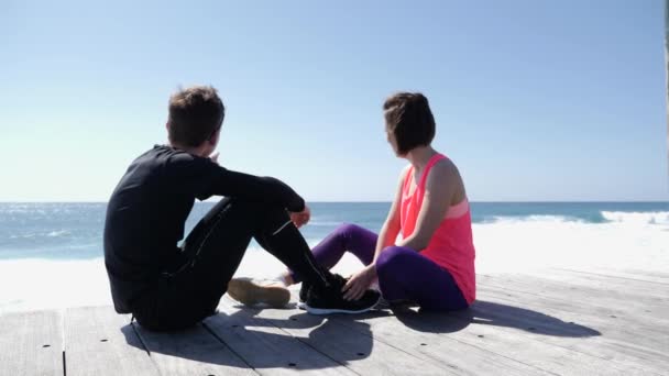Молодий чоловік і жінка сидять на кам'янистому пляжі. Хлопець вказує пальцем на море і небо і розмовляє — стокове відео
