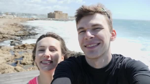 Joven feliz pareja amorosa sonriendo a la cámara en modo selfie en la playa rocosa. Fuertes olas golpeando las rocas . — Vídeo de stock