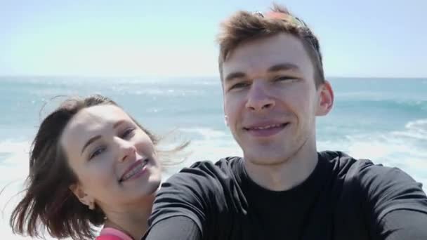 Jovem casal amoroso feliz sorrindo para a câmera no modo selfie na praia rochosa. Ondas fortes atingindo as rochas. Movimento lento — Vídeo de Stock