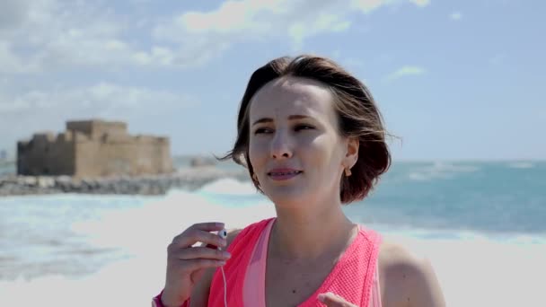 Joven hermosa mujer pone en los auriculares antes de entrenar en una playa. Fuertes olas están golpeando la costa. Movimiento lento — Vídeos de Stock