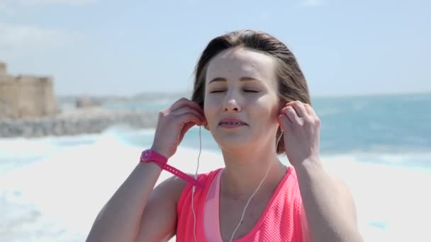 Fiatal kaukázusi Womans visel fejhallgatót a strandon. Erős szél és a hullámok. Vár a háttérben. Lassított mozgás — Stock videók