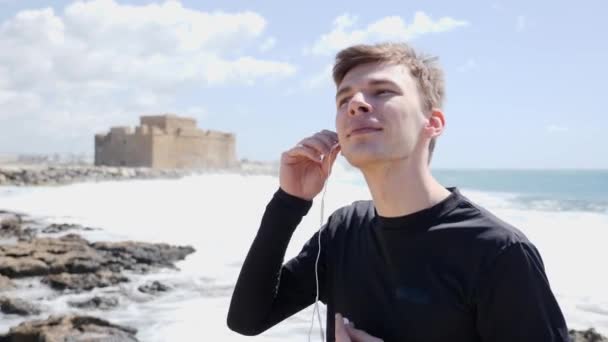 Joven hombre caucásico pone los auriculares en pie en la playa rocosa con el castillo en el fondo . — Vídeo de stock