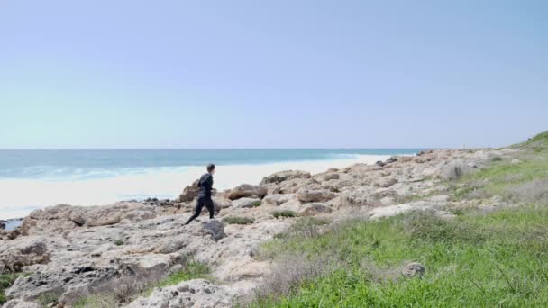Joven hombre caucásico en forma corriendo por un sendero rocoso a lo largo del océano. Vuelta atrás. Movimiento lento — Vídeo de stock