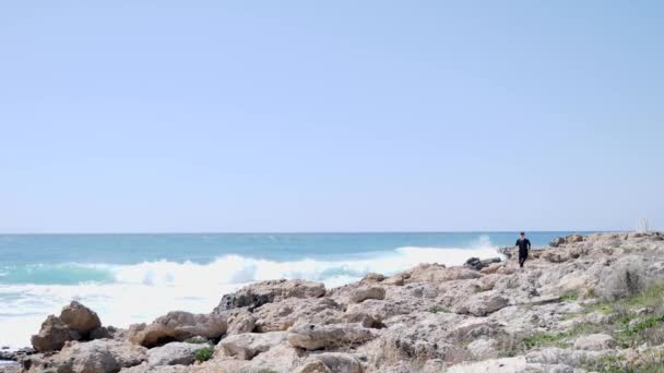 Homem atlético caucasiano correndo sobre as rochas em uma praia rochosa vestindo roupa preta . — Vídeo de Stock