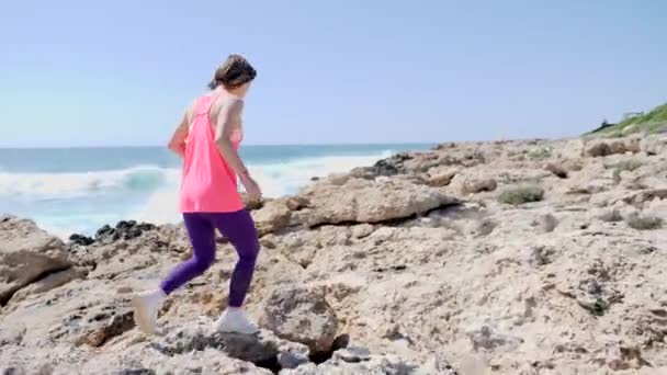 Joven chica deportiva corriendo por el camino rocoso vistiendo traje rosa. Vuelta atrás . — Vídeos de Stock