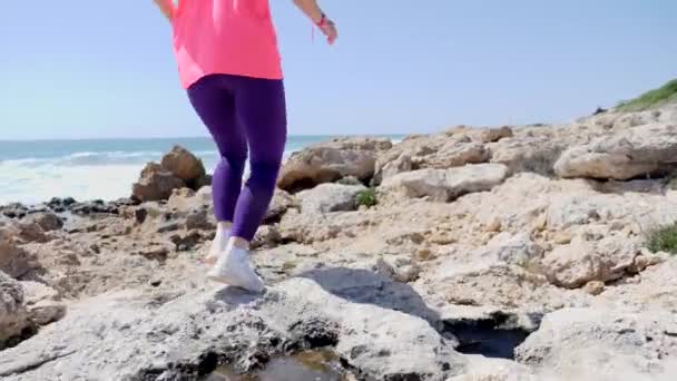 Mujer joven en forma corre el sendero rocoso a lo largo de la playa. El sendero corre sobre las rocas. A cámara lenta. Vuelta atrás — Vídeo de stock