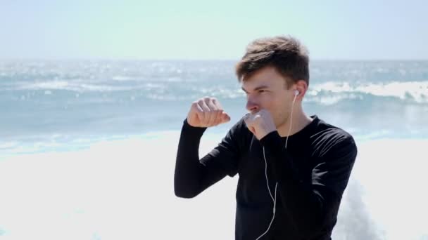 Jovem atleta caucasiano soca o ar usando fones de ouvido. Excertos de boxe na praia. Movimento lento — Vídeo de Stock