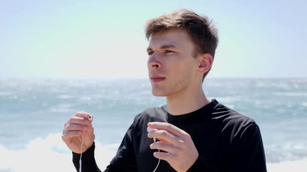 Jonge knappe man zet de koptelefoon op voor run workout staande op het strand — Stockvideo