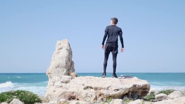 Joven hombre caucásico en forma de pie sobre la gran roca en la playa se pone sus gafas de sol y comienza a correr . — Vídeos de Stock