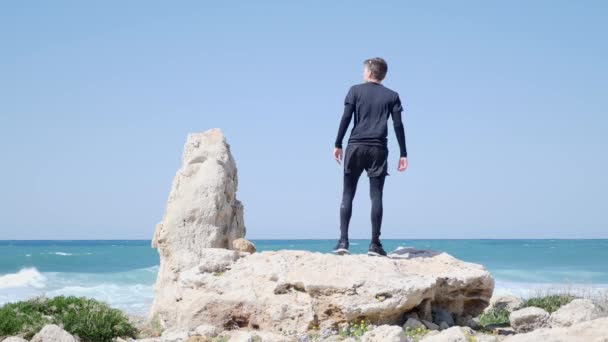 Junger fitter kaukasischer Mann, der auf dem großen Felsen am Strand steht, setzt seine Sonnenbrille auf und rennt los. — Stockvideo