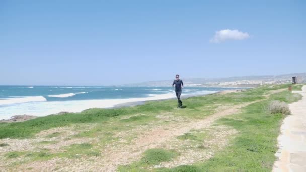Fiatal erős kaukázusi ember fut a strandon viselt fekete ruhában és napszemüveget. Ösvény fut végig a tengeren. Lassított mozgás — Stock videók