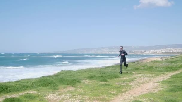 Joven hombre delgado caucásico corriendo en la playa con traje negro y gafas de sol. Día soleado ventoso — Vídeos de Stock