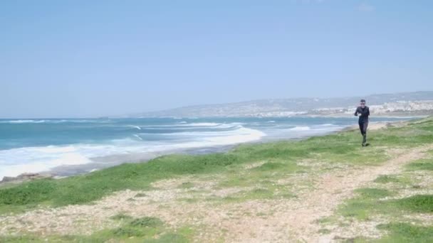 Jonge fit blanke man draait op het strand dragen zwarte outfit en zonnebrillen. Golvende zee is op de achtergrond — Stockvideo
