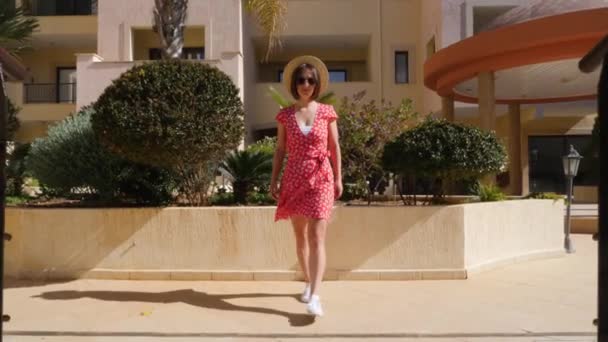Jeune femme caucasienne heureuse marchant sur le pont dans le jardin de l'hôtel vers la caméra en robe rouge, chapeau et lunettes — Video