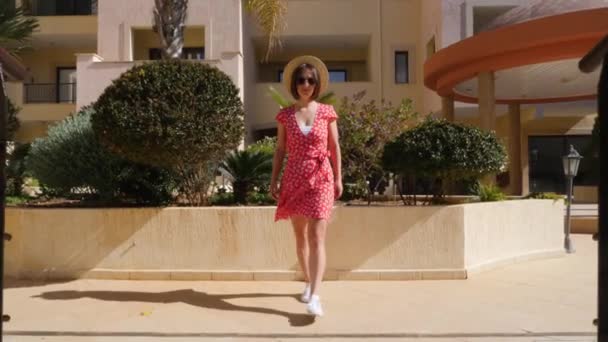 Young happy caucasian woman walking on the bridge in the hotel garden towards the camera wearing red dress, hat and glasses — 비디오