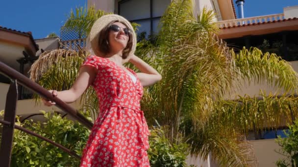 Jeune femme heureuse profite du soleil debout dans le jardin de l'hôtel avec des palmiers en robe rouge, chapeau et lunettes de soleil. Mouvement lent — Video