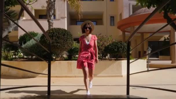Jeune femme séduisante en robe rouge avec lunettes de soleil et chapeau marchant sur le pont au-dessus de la piscine dans le jardin de l'hôtel palmier — Video