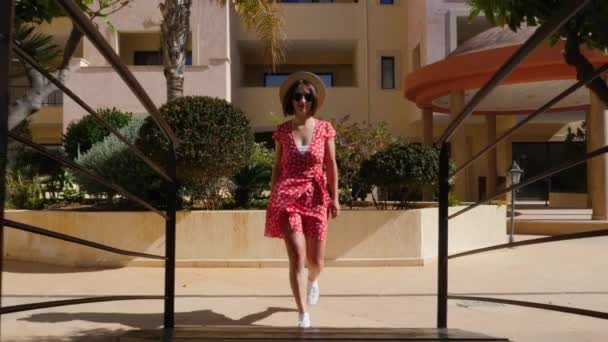 Junge attraktive Frau in rotem Kleid mit Sonnenbrille und Hut, die auf der Brücke über den Swimmingpool im Hotelgarten unter Palmen spaziert. Zeitlupe — Stockvideo