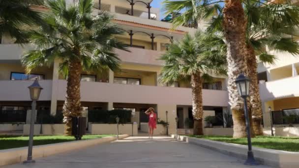 Attraktive schöne Frau, die im Hotelgarten in rotem Kleid, Hut und Sonnenbrille auf die Kamera zugeht und posiert. Palmen und Hotelgebäude. — Stockvideo