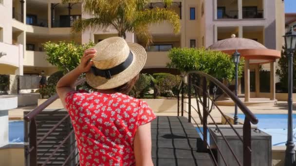 Νεαρή ευτυχισμένη γυναίκα προσκαλεί τον άνδρα να την ακολουθήσει με το χέρι της φορώντας κόκκινο φόρεμα, γυαλιά ηλίου και καπέλο. Γέφυρα πάνω από την πισίνα στον κήπο του ξενοδοχείου με φοίνικες. Αργή κίνηση — Αρχείο Βίντεο