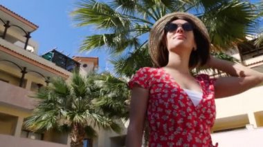 Güneşten hoşlanan, arka planda palmiye ağaçları olan şapka ve güneş gözlüğü takan genç ve çekici bir kadın. Otel bahçesi. Yavaş çekim