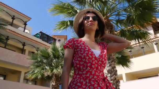 Młoda atrakcyjna kobieta ciesząca się słońcem i pozująca w kapeluszu i okularach przeciwsłonecznych z palmami na tle. Hotelowy ogród — Wideo stockowe