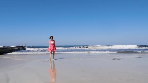 Jovem mulher atraente caminhando em direção à câmera na praia desfrutando do sol e do vento. Mar cais céu azul e ondas tempestuosas no fundo. Movimento lento — Vídeo de Stock