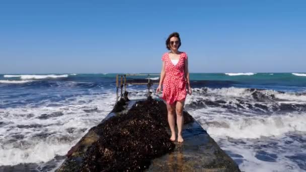 Joven mujer feliz vistiendo vestido rojo y gafas de sol de pie en el muelle con olas de mar golpeando contra el muelle . — Vídeos de Stock