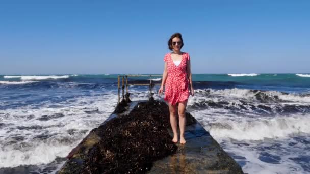 Joven mujer feliz vistiendo vestido rojo y gafas de sol de pie en el muelle con olas de mar golpeando contra el muelle. Movimiento lento — Vídeo de stock