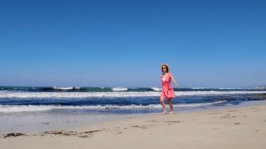 Genç beyaz kadın kumsalda yalınayak koşuyor ve şapkasını gökyüzüne fırlatıyor. Kırmızı elbise ve arka planda güçlü dalgalar olan güneş gözlüğü takıyor. Yavaş çekim