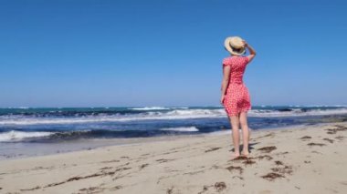 Genç çekici bir kadın şapkasını tutuyor ve kırmızı elbisesiyle fırtınalı denize ve ufka bakıyor. Kırmızı elbise rüzgarda çırpınıyor..
