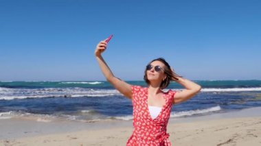 Kırmızı elbiseli genç bir kadın, deniz sahilinde güçlü rüzgar ve dalgalarla fotoğraf çekiyor. Yavaş çekim