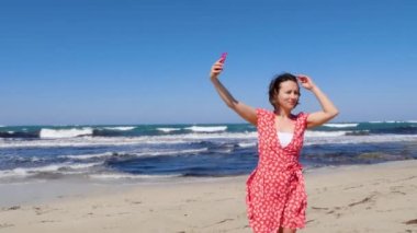 Genç ve tatlı bir kadın güneş gözlüğünü takıp arka planda güçlü dalgalarla kumsalda dururken selfie çekiyor..