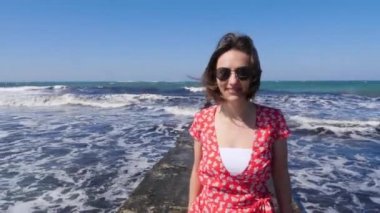 Gülümseyen ve iskelede yürüyen genç, tatlı bir kadın. Güçlü dalgalar arka plana vuruyor ve sıçrıyor..