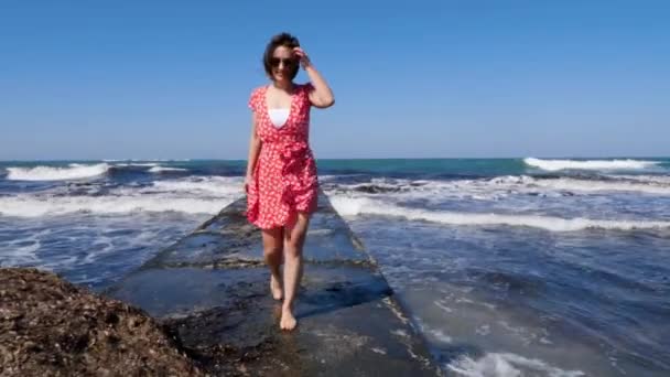 Joven mujer feliz sonriente caminando sobre un muelle de mar descalzo. Vestido rojo aleteando en el viento. Olas golpeando el muelle . — Vídeos de Stock