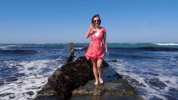 Mujer atractiva joven caminando descalza en el muelle del mar hacia la cámara en vestido rojo y gafas de sol. Olas tormentosas golpeando el muelle. Movimiento lento — Vídeos de Stock