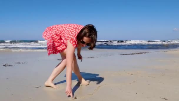 Young happy lady draws pictures on the sea beach sand. Strong stormy waves hitting the seashore. Wind blows. Slow motion — Stock Video