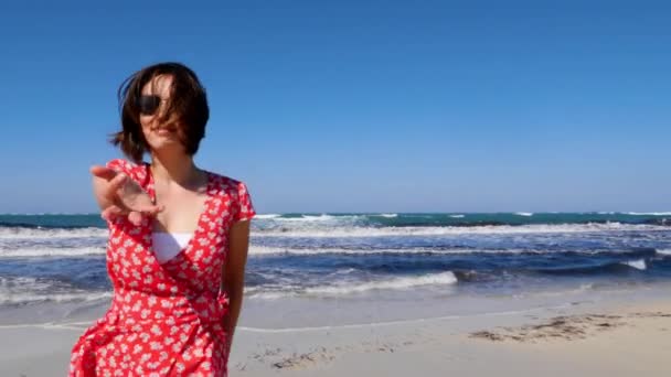 Νεαρή χαμογελαστή γυναίκα καλεί έναν άντρα να την ακολουθήσει περπατώντας προς τη θάλασσα. Δυνατά κύματα χτυπούν την παραλία. Κόκκινο φόρεμα και γυαλιά ηλίου. Αργή κίνηση — Αρχείο Βίντεο