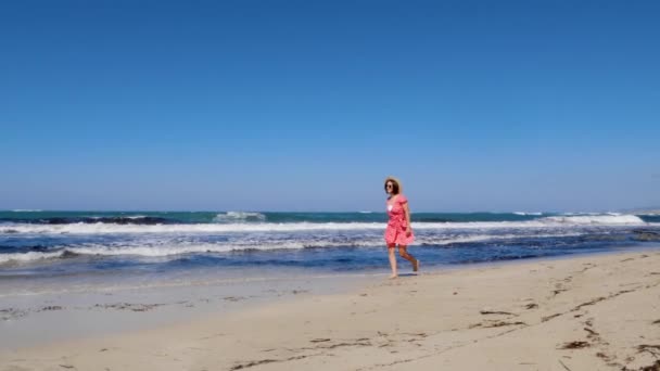 Jovem mulher alegre caminha ao longo da praia e joga seu chapéu fora vestindo vestido vermelho e óculos de sol. Fortes ondas tempestuosas e céu azul no fundo. Movimento lento — Vídeo de Stock