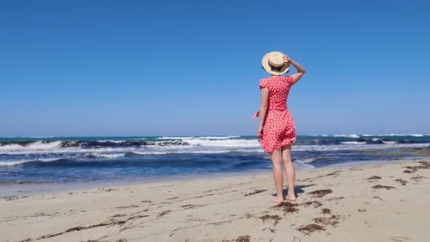 Genç ve çekici bir kadın şapkasını tutarken kırmızı elbise güçlü rüzgarda kanat çırpıyor. Boş sahilde durur ufka ve deniz dalgalarına doğru bakar.. — Stok video