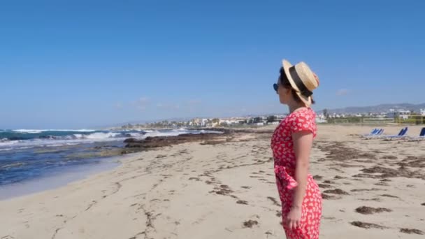 Fiatal gyönyörű nő piros ruhában és kalapban áll egyedül az üres strandon napágyakkal. Homokos strand erős hullámokkal és széllel. — Stock videók