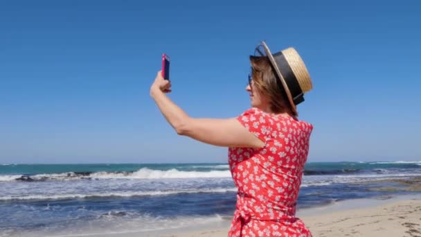 Young woman holding her phone and taking pictures of the windy stormy sea and waves. She wears hat and red dress with sunglasses. — Stock Video