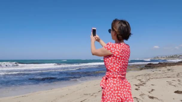 Mujer caucásica sosteniendo el teléfono de la cámara y tomando fotos de la tormenta marina de pie en la playa. Lleva vestido rojo y gafas de sol. . — Vídeos de Stock